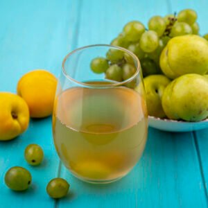 side view of white grape juice in glass and fruits as grape and green pluots in bowl with nectacots and grape berries on blue background