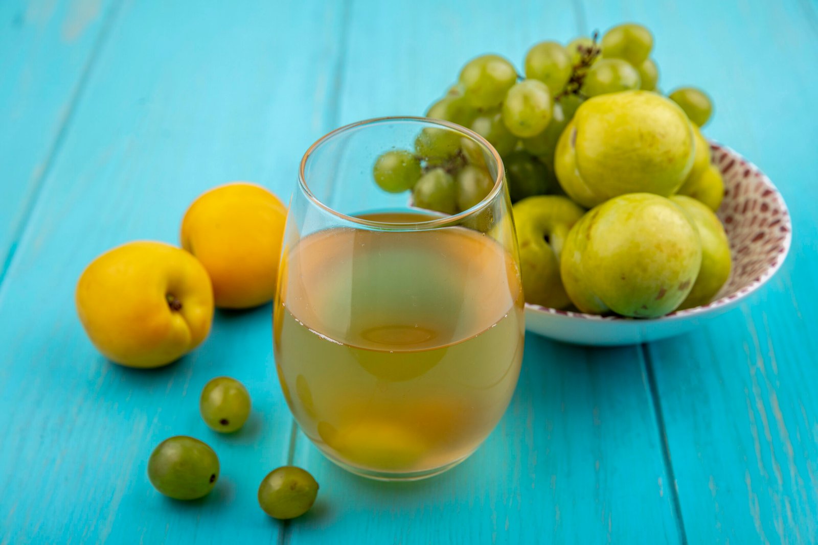 side view of white grape juice in glass and fruits as grape and green pluots in bowl with nectacots and grape berries on blue background side view of white grape juice in glass and fruits as grape and green pluots in bowl with nectacots and grape berries on blue background